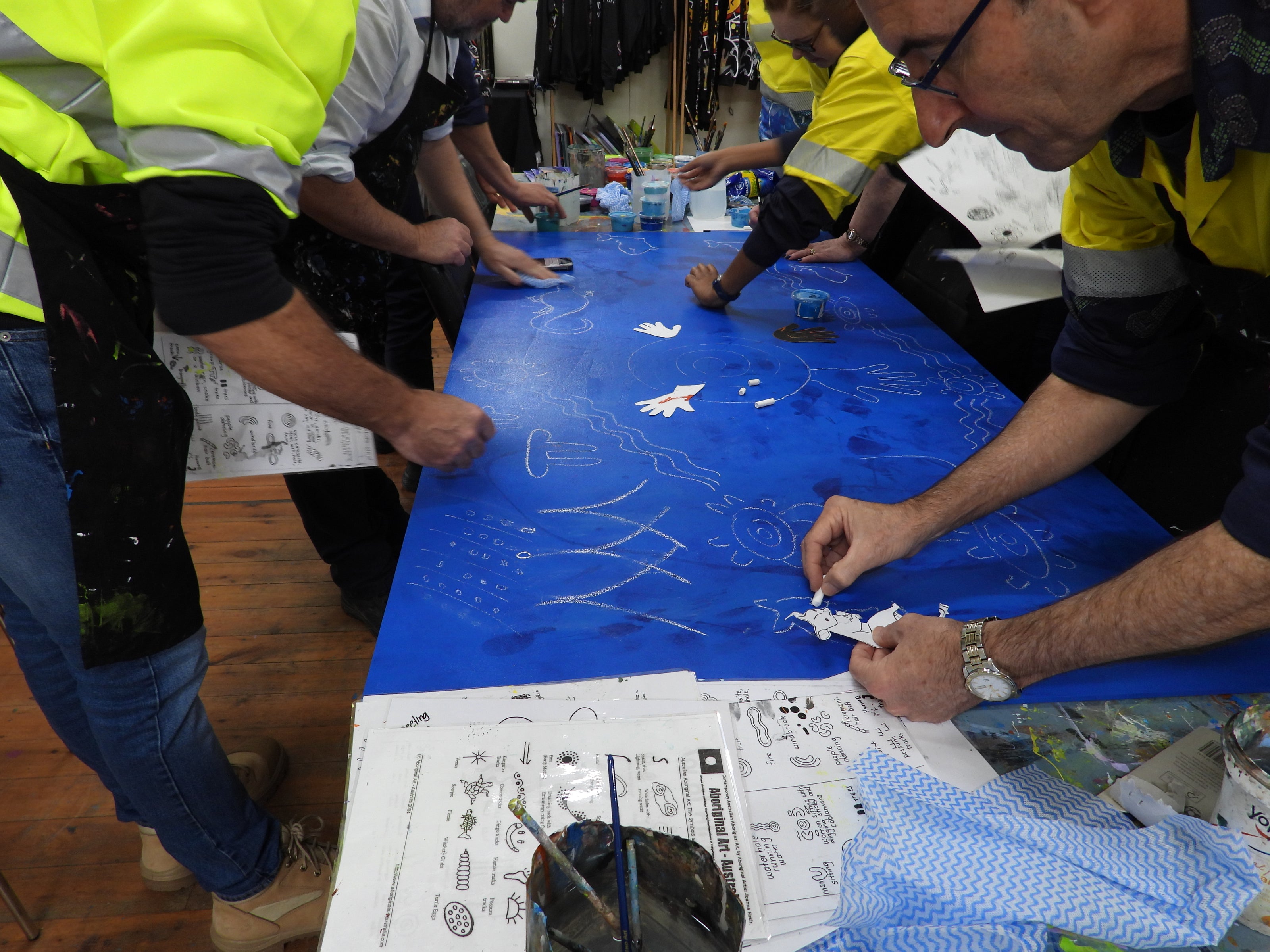 Group of people working on a large blue painting with various art supplies around.